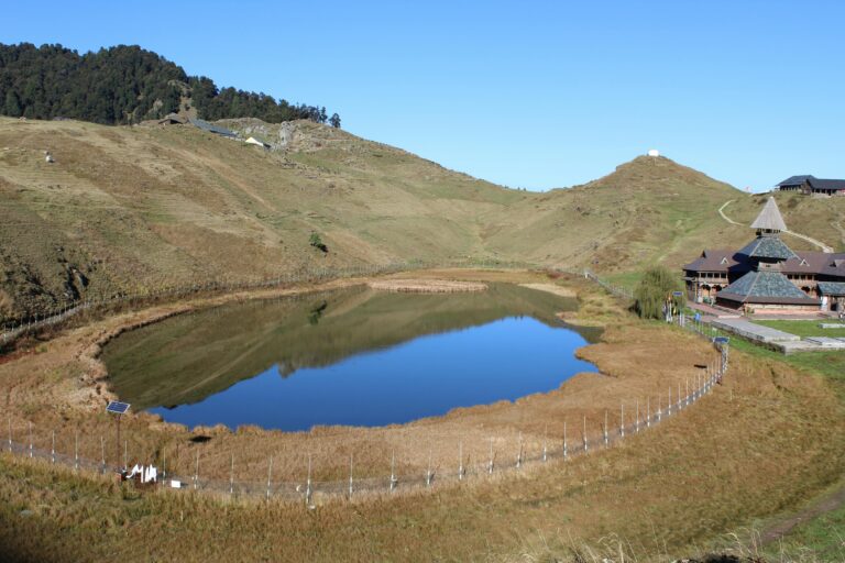 A breathtaking view of Prashar Lake surrounded by lush green meadows and snow-capped mountains, with a small floating island in the center and the historic Prashar Rishi temple nearby.